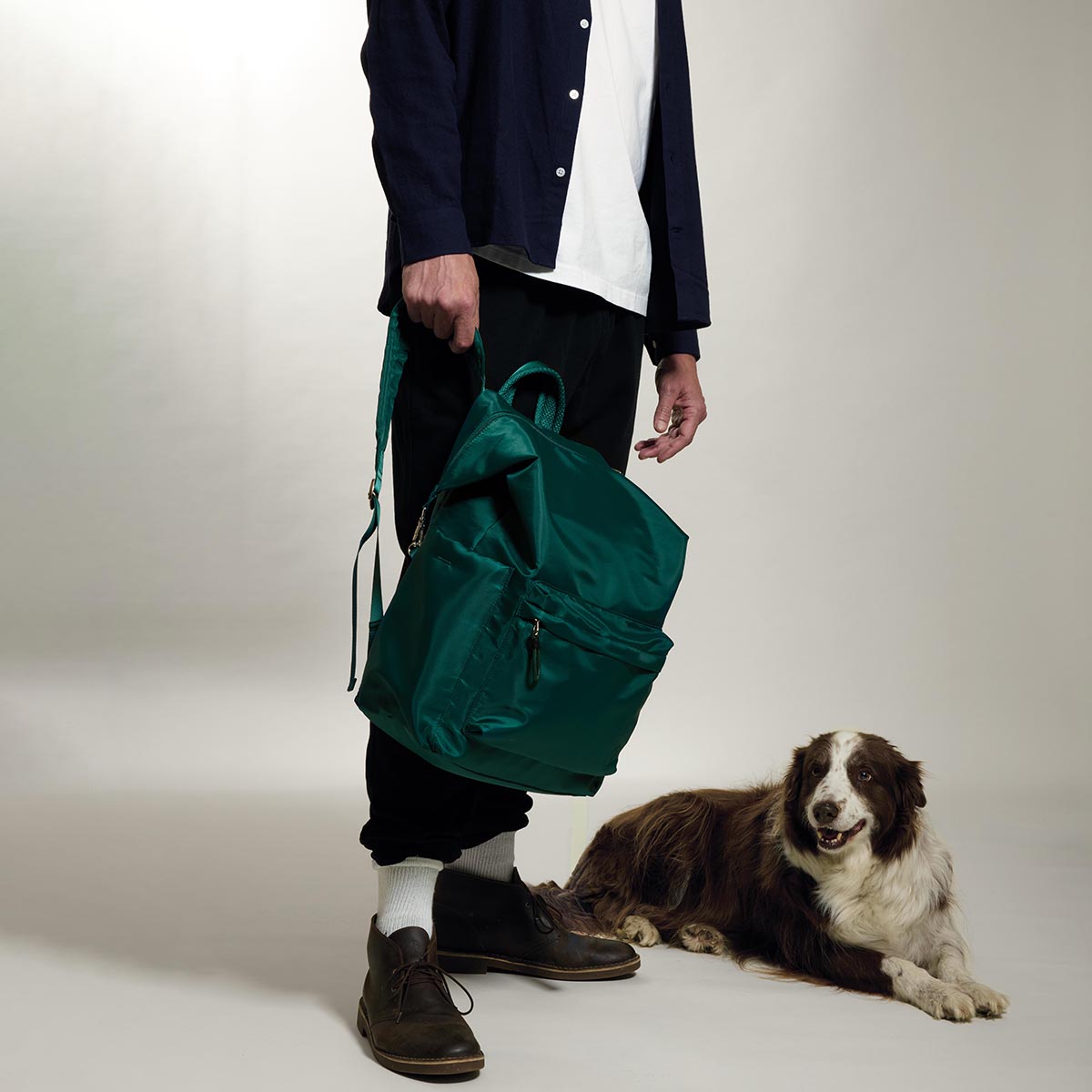Practice The Karma Backpack in Forest green color held by model wearing black pants by striped long haired dog
